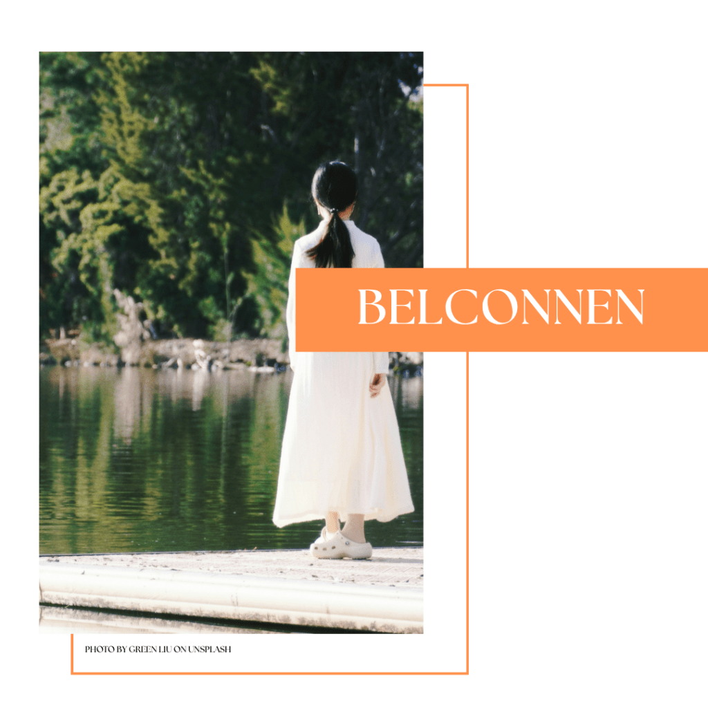 A person wearing a long white dress and clogs stands on a dock by a lake, surrounded by greenery.